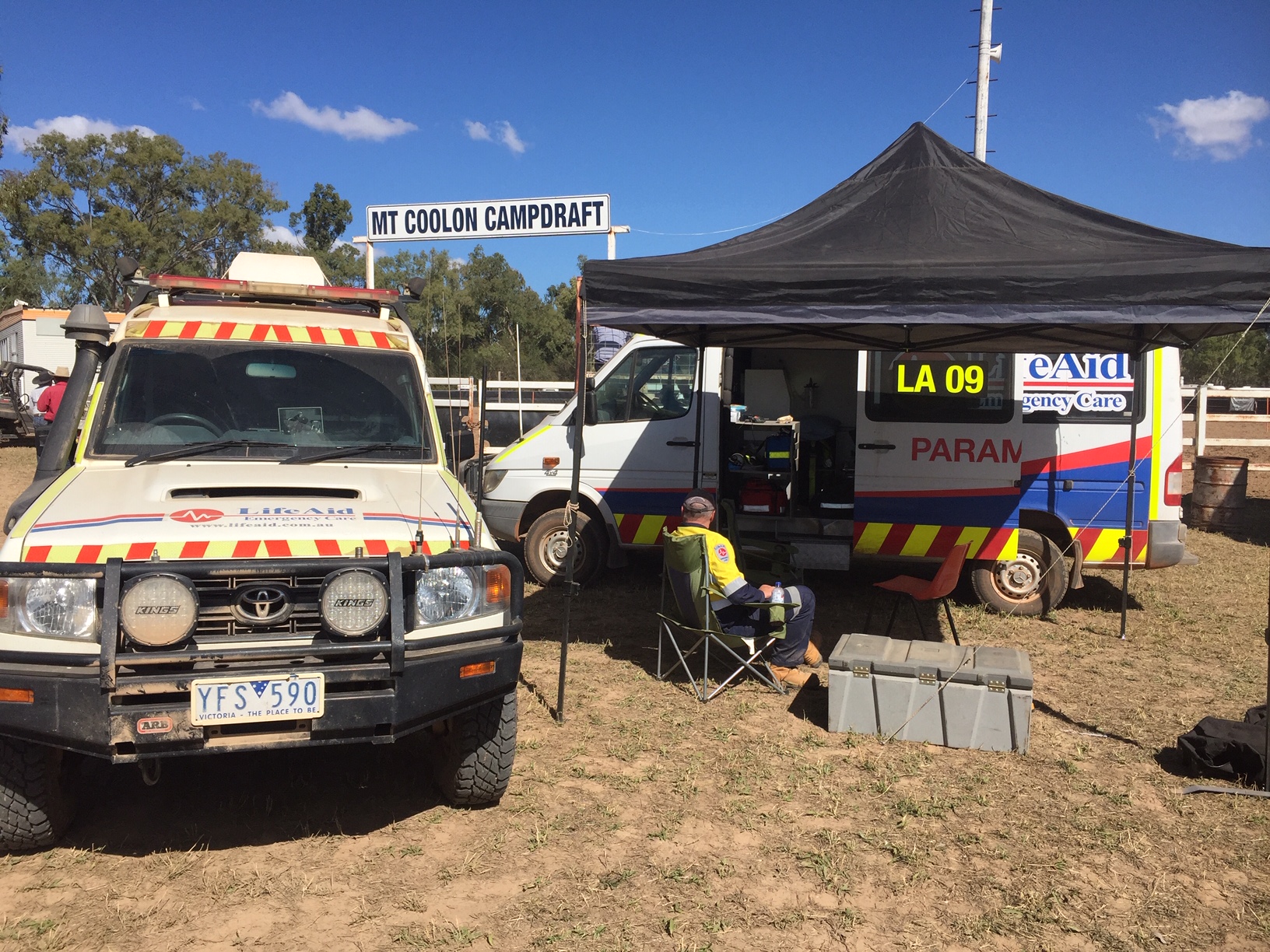 Two 4WD ambulances with the LifeAid logo, and a paramedic sitting in a camp chair in the shade beside them.